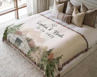 May include: A beige woven blanket with a watercolor mountain and tent design. The blanket features the names "Sophia & Noah" and the date "10-28-2025" in black script. The blanket has a fringed edge and is displayed on a bed with decorative pillows.