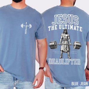 May include: A light blue t-shirt with a white graphic of a barbell and cross on the front. The back of the shirt features the text "JESUS THE ULTIMATE DEADLIFTER" with an illustration of Jesus lifting weights. The words "BLUE JEAN" are at the bottom.