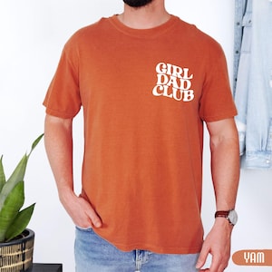 Op de afbeelding: Oranje-bruin T-shirt met korte mouwen en de witte tekst "GIRL DAD CLUB" verticaal op de borst. Het shirt is effen van kleur en lijkt gemaakt van een zacht, comfortabel materiaal.