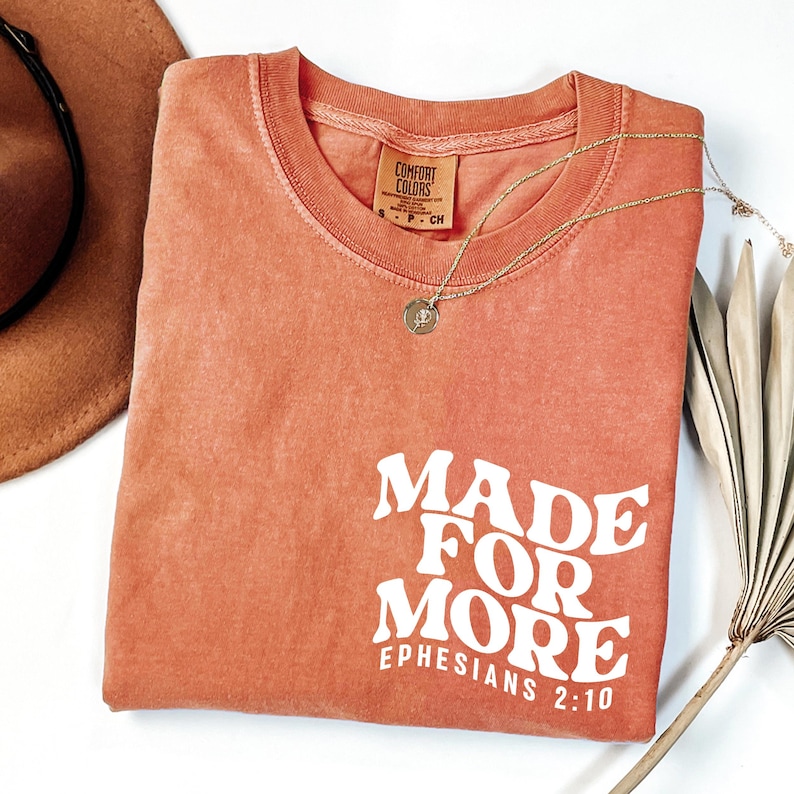 May include: A coral-colored t-shirt with the phrase "MADE FOR MORE" and "Ephesians 2:10" printed in white. A gold necklace with a pendant and a brown hat are visible. The shirt has a crew neck and short sleeves.