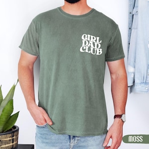 Op de afbeelding: Mosgroen t-shirt met de witte tekst "GIRL DAD CLUB" op de linkerborst. Het shirt met korte mouwen is effen van kleur. Het model draagt een blauwe spijkerbroek en een horloge met een bruine leren band.
