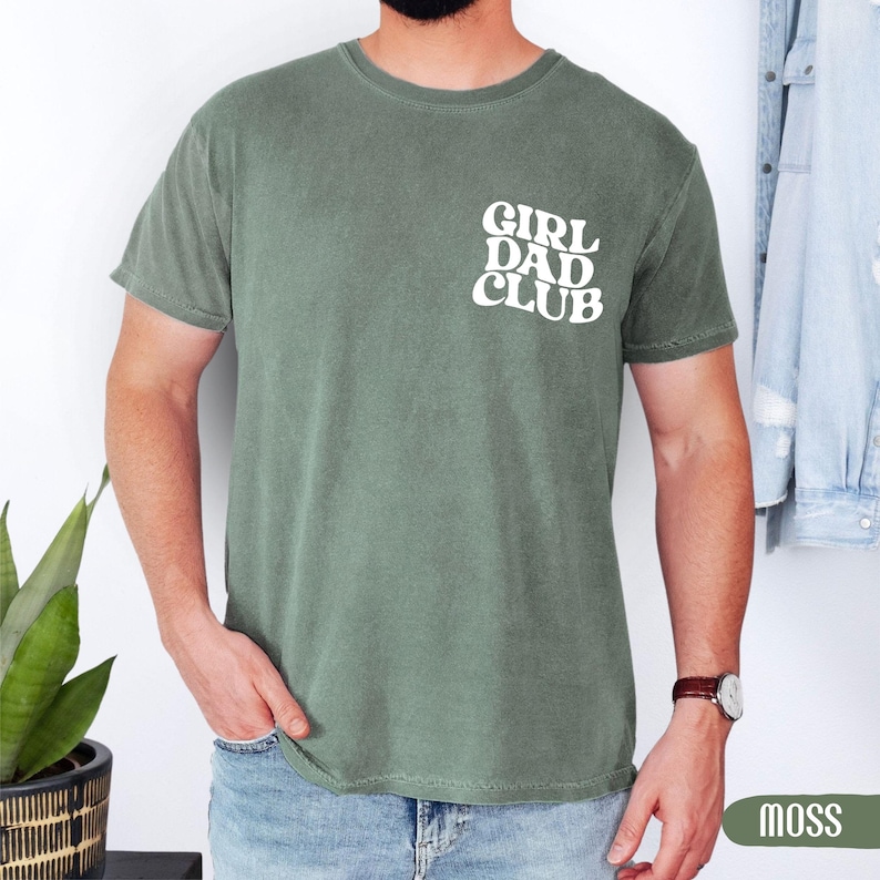 Op de afbeelding: Mosgroen t-shirt met de witte tekst "GIRL DAD CLUB" op de linkerborst. Het shirt met korte mouwen is effen van kleur. Het model draagt een blauwe spijkerbroek en een horloge met een bruine leren band.