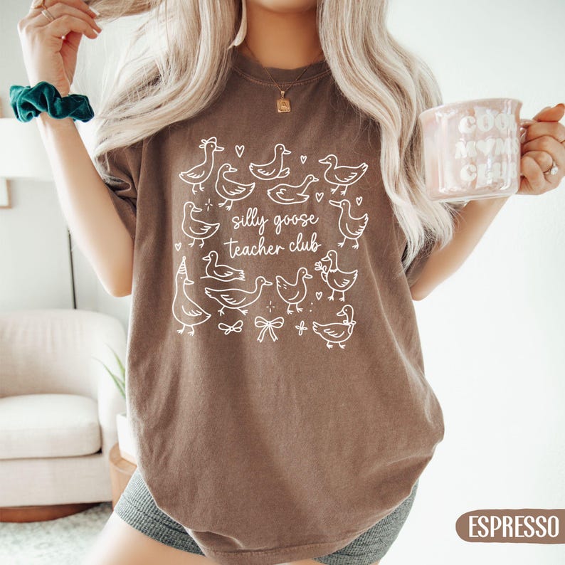 Puede incluir: Camiseta color espresso con un gr&aacute;fico blanco de gansos de dibujos animados y el texto "silly goose teacher club". La camiseta es de cuello redondo y tiene mangas cortas. Una taza con texto se sostiene en la mano.