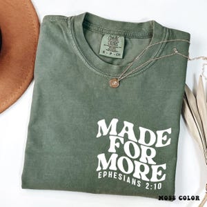 May include: A moss green t-shirt with the text "MADE FOR MORE EPHESIANS 2:10" in white. The shirt has a crew neck and short sleeves. A gold necklace with a pendant is draped across the shirt. The words "MOSS COLOR" are at the bottom.