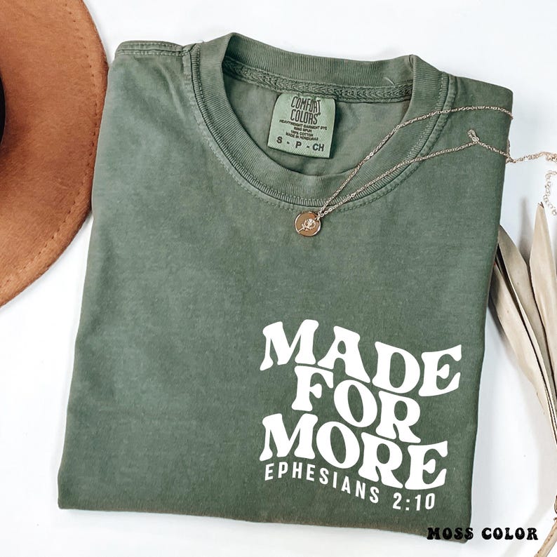 May include: A moss green t-shirt with the text "MADE FOR MORE EPHESIANS 2:10" in white. The shirt has a crew neck and short sleeves. A gold necklace with a pendant is draped across the shirt. The words "MOSS COLOR" are at the bottom.