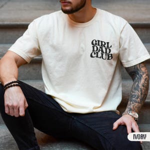 Op de afbeelding: Ivoor kleurig T-shirt met de tekst "GIRL DAD CLUB" in zwart, met een ronde hals en korte mouwen. Het shirt is gemaakt van een zacht materiaal. Het woord "IVORY" staat rechtsonder.