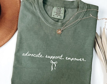 May include: A moss green t-shirt with the words "advocate. support. empower." in white script. A gold necklace with a pendant rests on the shirt. The shirt is folded and placed next to a brown hat.
