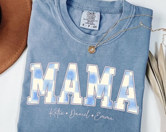 Op de afbeelding: Een blauw Comfort Colors T-shirt met het woord "MAMA" in grote, witte letters met een blauw-wit geblokt patroon. Onder het woord "MAMA" staan de namen "Katie", "Daniel" en "Emma". Een gouden ketting ligt over het shirt.