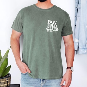 May include: Olive green t-shirt with the text "BOY DAD CLUB EST. 2025" printed in white on the upper left chest. The shirt is a crew neck style and has short sleeves. The model is wearing blue jeans and a brown watch.
