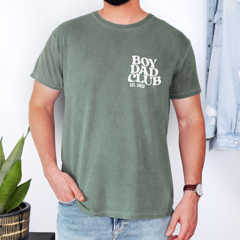 May include: Olive green t-shirt with the text "BOY DAD CLUB EST. 2025" printed in white on the upper left chest. The shirt is a crew neck style and has short sleeves. The model is wearing blue jeans and a brown watch.