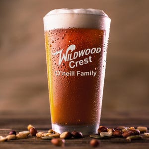 Peut inclure: Un grand verre de bi&egrave;re ambr&eacute;e avec une &eacute;paisse mousse blanche. Le verre est couvert de condensation et porte l'inscription "Wildwood Crest O'Neill Family". Des noix m&eacute;lang&eacute;es sont dispers&eacute;es sur une surface en bois.