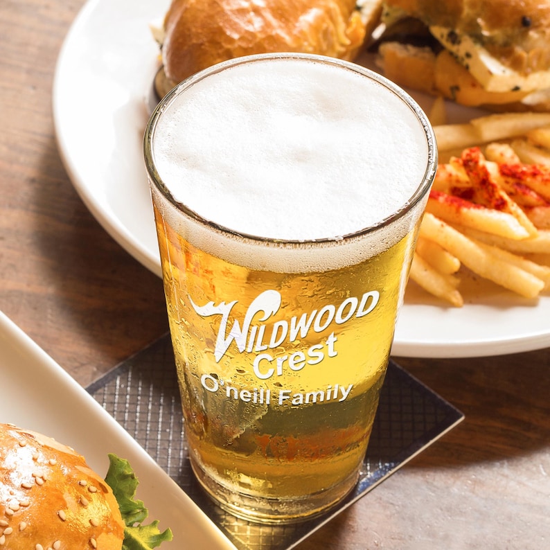 Peut inclure: Un verre transparent rempli de bi&egrave;re et surmont&eacute; d'une mousse blanche. Le verre porte l'inscription "Wildwood Crest O'Neill Family". Le verre est pos&eacute; sur un dessous de verre, avec des hamburgers et des frites en arri&egrave;re-plan.