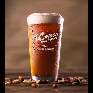 May include: A frosted glass beer mug with a white logo that reads "Wildwood New Jersey The Lynch Family". The mug is filled with a dark amber beer and condensation on the outside of the glass.