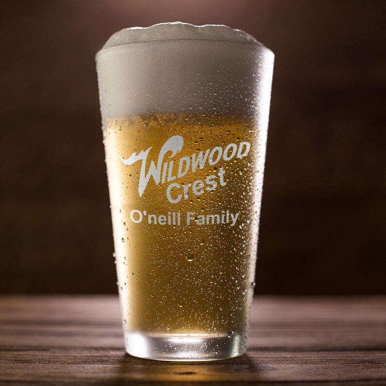 Peut inclure: Un verre transparent rempli de bi&egrave;re blonde et surmont&eacute; d'une &eacute;paisse mousse blanche. Le verre porte les inscriptions blanches "Wildwood Crest" et "O'Neill Family". Il est couvert de condensation et pos&eacute; sur une surface en bois.