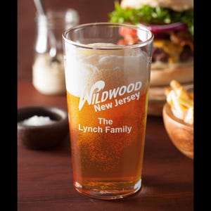 May include: A clear glass beer mug with a white inscription that reads "Wildwood New Jersey The Lynch Family". The mug is filled with a light amber beer and has a white foam head.