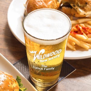 May include: A frosted glass beer mug with the text "Wildwood New Jersey The Lynch Family" printed on the side. The mug is filled with a light yellow beer and is sitting on a brown wooden surface.