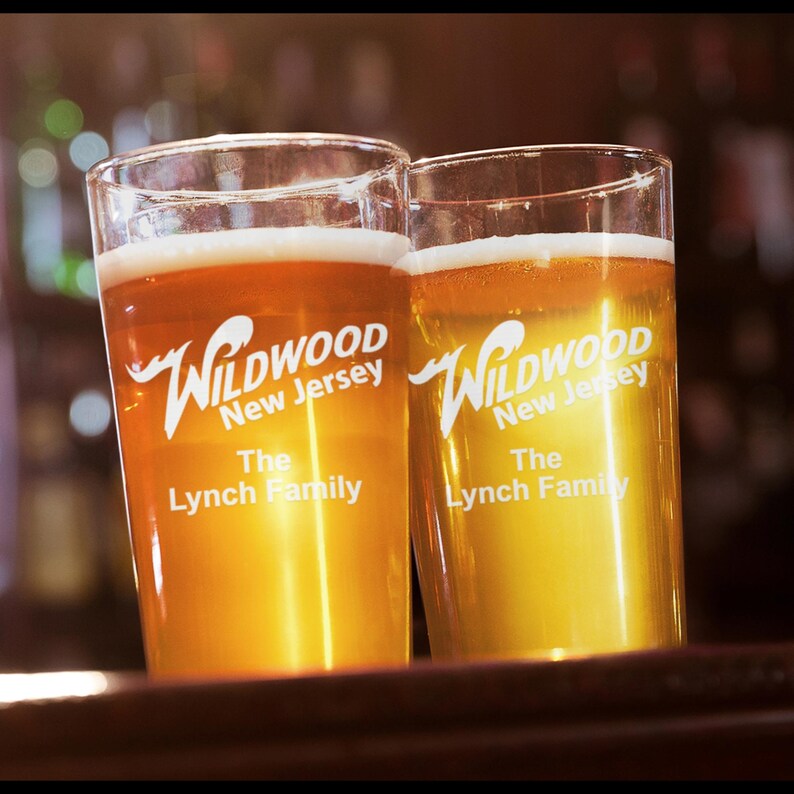 May include: Two clear glass pint glasses filled with a light amber colored beer. Both glasses have the text "Wildwood New Jersey The Lynch Family" printed on them.