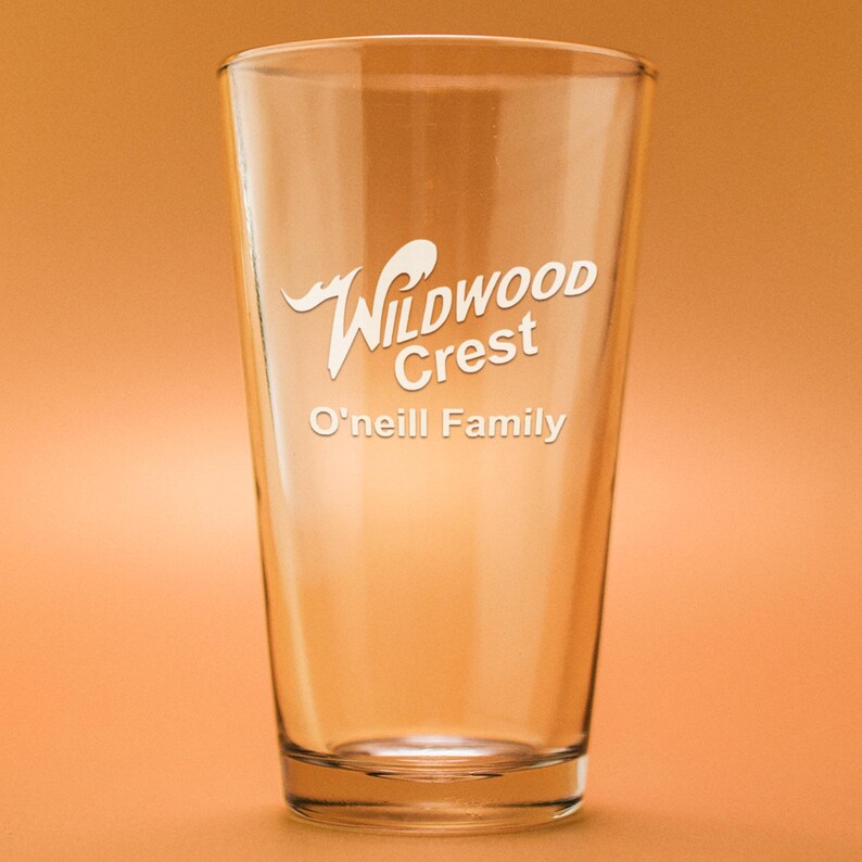 Peut inclure: Un verre &agrave; bi&egrave;re transparent avec les mots "Wildwood Crest" et "O'neill Family" en &eacute;criture blanche. Le verre est vide et se trouve sur un fond orange.