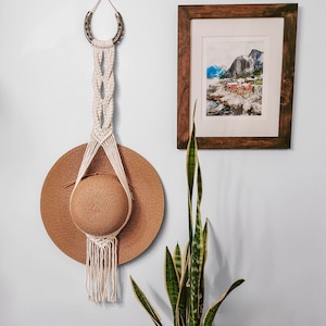 May include: A white macrame wall hanging with a brown woven hat hanging from it. The macrame is made with a simple knot pattern and has a horseshoe-shaped top. The hat is a wide-brimmed straw hat with a brown band.