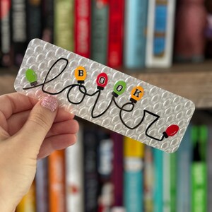 Puede incluir: Un marcapáginas blanco con un contorno negro de la palabra "lover" con una cadena de luces navideñas que deletrea "BOOK" en el medio. Las luces son rojas, verdes, amarillas y naranjas.