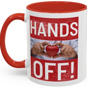 May include: White ceramic mug with a red rim and handle. The mug has a red background with white text that says "HANDS OFF!" and a red heart in the center. The heart is being held by two hands.