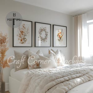 May include: Three framed watercolor prints of floral arrangements hang above a white bed with a white duvet and several pillows. The prints are in black frames and feature a wreath, a bouquet of flowers, and a cluster of wildflowers.
