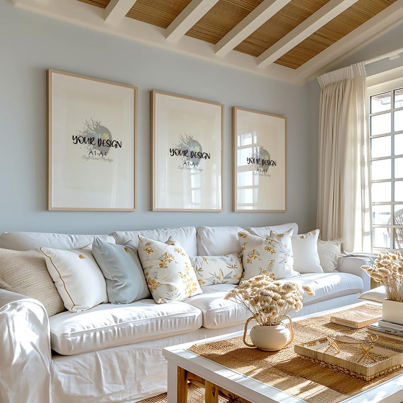 May include: Three framed prints with the text "YOUR DESIGN ATLAS" hanging on a light blue wall above a white couch with pillows. The couch is in a living room with a coffee table and dried flowers in a vase.