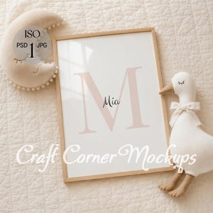 May include: A rectangular wooden frame containing a white print with a large pale pink letter M and the name Mia in black script. A beige crescent moon plush with pom-pom trim and a white plush goose with a bow tie are positioned nearby.