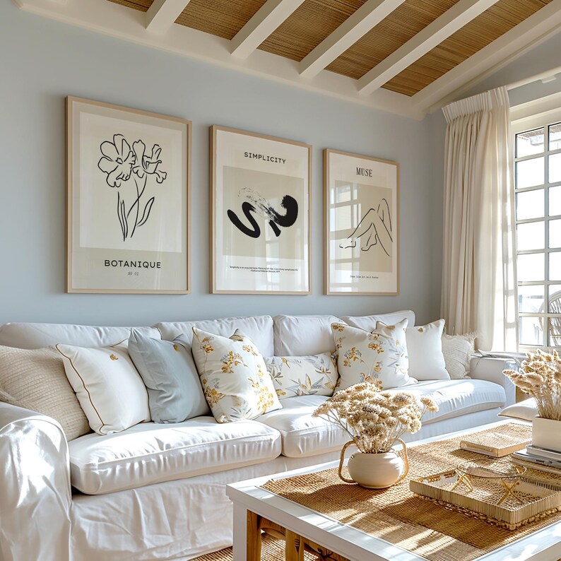May include: A white couch with a white throw blanket and several throw pillows in a living room with a white coffee table and a vase of dried flowers. Three framed prints hang on the wall behind the couch. The prints feature black and white line drawings of a flower, an abstract shape, and a woman's legs. The text on the prints reads "Botanique", "Simplicity", and "Muse".