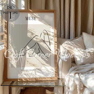 Op de afbeelding: Een ingelijst print met een minimalistisch line art-ontwerp van een zittend persoon met het woord "MUSE" boven de afbeelding. De print is in een houten lijst en staat op een glazen tafel voor een beige bank.