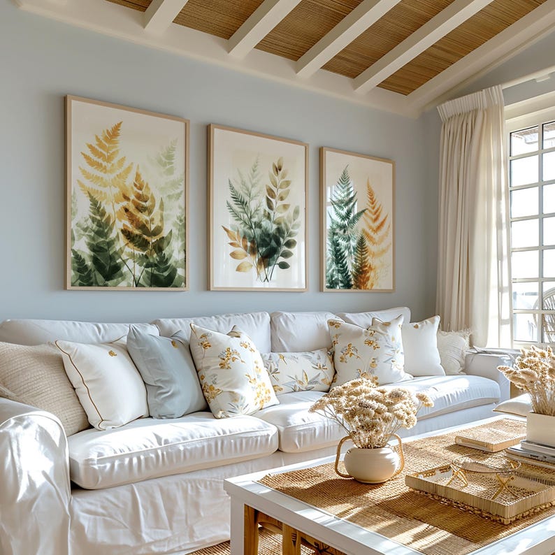 May include: A white couch with a variety of throw pillows in a living room setting. The couch is in front of a wall with three framed prints of watercolor fern leaves in shades of green and brown. The room has a light blue wall color and a wooden ceiling.