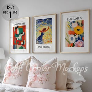 May include: Three framed art prints with gold frames and white mats. The prints feature colorful abstract paintings by Henri Matisse. The prints are hanging on a white wall above a bed with white bedding and pink floral throw pillows.