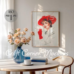 May include: A framed print of a woman wearing a red hat and a white coat, holding a red glove. The print is hanging on a white wall above a wooden table with a blue glass vase filled with beige flowers, a stack of books, and a white coffee mug.