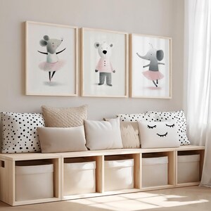 May include: Three framed wall art prints featuring watercolor illustrations of a mouse in a pink tutu, a bear in a striped shirt, and an elephant in a pink tutu, displayed above a light wood storage bench with five fabric bins and pillows.