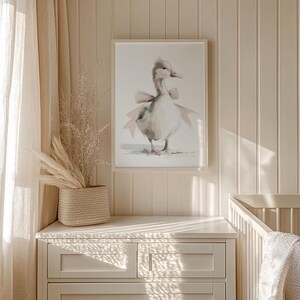 May include: A framed watercolor print of a goose wearing a pink bow. The artwork is displayed on a white dresser in a room with beige walls and a woven basket with dried plants. A crib is visible in the background.