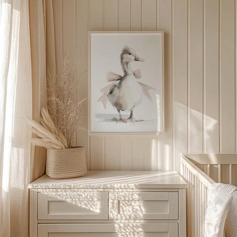 May include: A framed watercolor print of a goose wearing a pink bow. The artwork is displayed on a white dresser in a room with beige walls and a woven basket with dried plants. A crib is visible in the background.