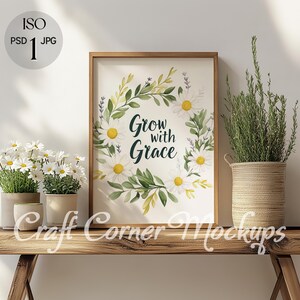 May include: A framed print with a white background and a floral wreath with white daisies and green leaves. The wreath surrounds the text "Grow with Grace". The print is sitting on a wooden shelf with two potted plants.