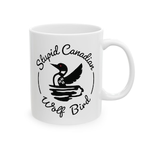 Pode incluir: Caneca de cerâmica branca com um gráfico preto de um mergulhão e o texto "Stupid Canadian Wolf Bird" em um design circular. O pássaro tem um olho vermelho e é retratado na água.