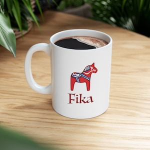 May include: White ceramic mug with a red and blue Dala horse illustration and the word "Fika" in red lettering. The mug is filled with black coffee.
