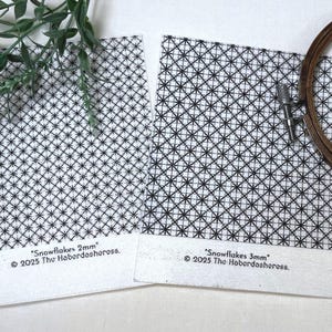 Pode incluir: Dois quadrados de tecido branco com padrões de flocos de neve pretos, rotulados como "Snowflakes 2mm" e "Snowflakes 3mm". Também estão presentes um bastidor de bordado de madeira e vegetação. O texto "© 2025 The Haberdasheress" está impresso no tecido.