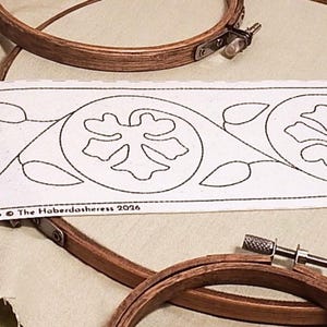 Op de afbeelding: Een witte stoffen strook met een zwart lijnpatroon van herhalende cirkelvormige bloemmotieven en bladeren. De tekst London 8cm BB048Lo 8cm The Haberdasheress 2026 staat onderaan gedrukt. Twee houten borduurringen zijn zichtbaar.