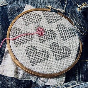 May include: A close-up of a wooden embroidery hoop with a white fabric stretched across it. The fabric is embroidered with a pattern of five hearts, each made up of small squares. The hearts are stitched in a dark gray color. The hoop is sitting on a piece of blue denim fabric.