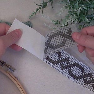 Op de afbeelding: Een ambachtelijk project in uitvoering, met een houten borduurring, groen garen en een zelfklevende strip met patroon. De strip heeft een geometrisch ontwerp in grijs en wit, met de tekst "The Haberdasheress 2025".