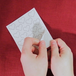 Op de afbeelding: Een wit vel papier met een raster van getekende harten. Het vel wordt van een achterblad gepeld.