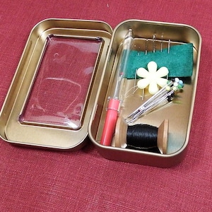 Op de afbeelding: Een gouden metalen doos met een transparant plastic venster. De doos bevat een rood en doorzichtig naaiwerktuig, een spoel zwart garen, een houten spoel en een groen viltkussen met gele bloemspelden.