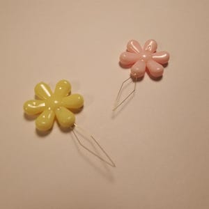 May include: Two flower-shaped threaders, one pink and one yellow, with a thin metal wire attached to each.
