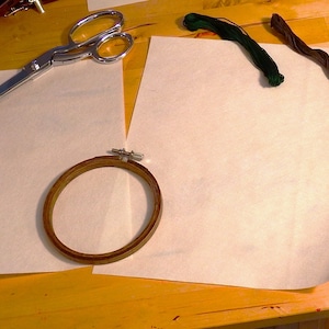 Op de afbeelding: Een houten borduurring met een witte stof erover gespannen, een zilveren schaar en twee spoelen borduurdraad, een groene en een zwarte, op een houten oppervlak.