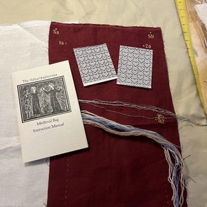 May include: A medieval bag craft kit. It includes a burgundy fabric piece, two cards with heart patterns, and a booklet titled "Medieval Bag Instruction Manual." Embroidery floss in various colours and a white fabric piece are also present.