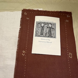 May include: A medieval bag instruction manual with a black and white illustration of two figures. The manual is on a brown fabric with dotted lines and numbers. A piece of white fabric is to the left.