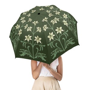 Può includere: Un ombrello aperto con sfondo verde scuro e motivo floreale color crema e verde chiaro. Il design dell'ombrello presenta fiori e foglie stilizzate, creando un aspetto decorativo ed elegante. L'ombrello è aperto.